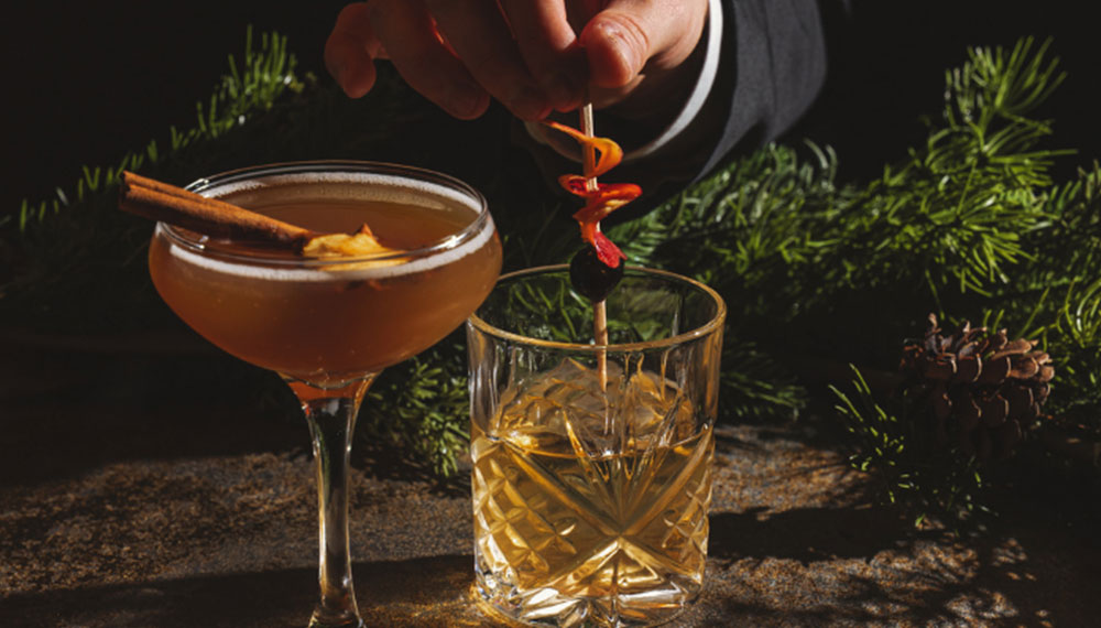 Two winter cocktails on a table with evergreen décor—one garnished with a cinnamon stick, the other with a citrus peel and cherry held by a hand.